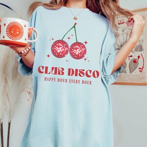 May include: Light blue t-shirt with a red cherry graphic. The cherry is made of disco balls. The text "Club Disco" and "Happy Hour Every Hour" is printed in red.