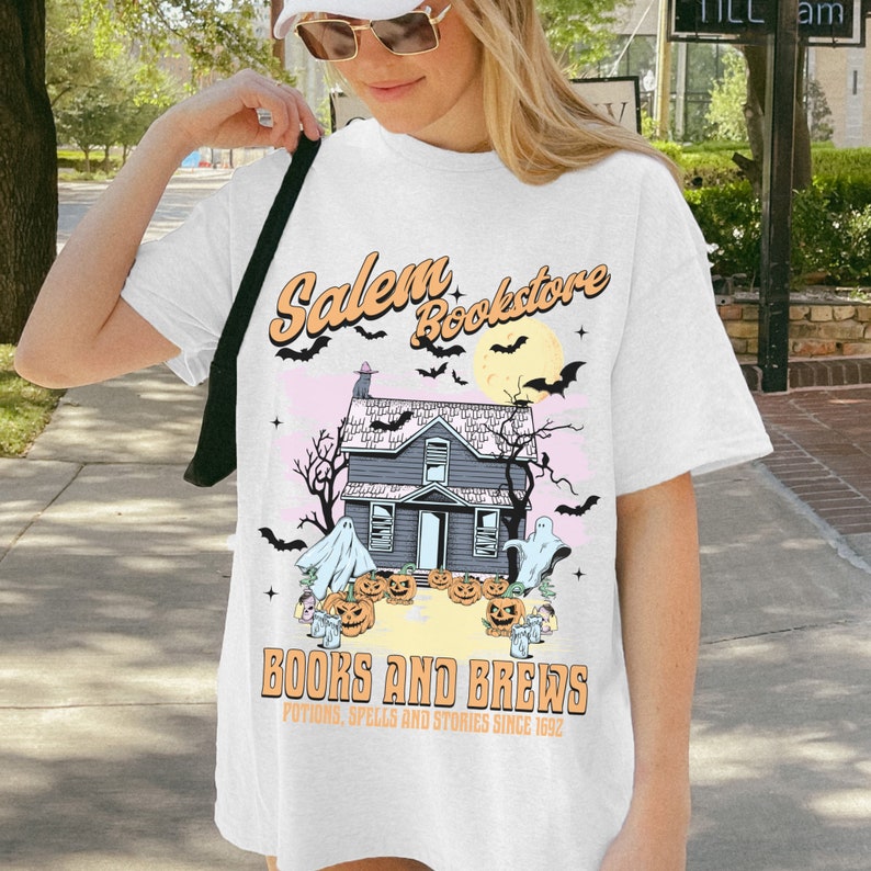 Op de afbeelding: Een wit T-shirt met een grafisch ontwerp van een spookhuis met pompoenen en geesten. De tekst "Salem Bookstore" staat boven het huis en "Books and Brews" staat onder het huis. De tekst "Potions, Spells and Stories Since 1692" staat onder het huis.