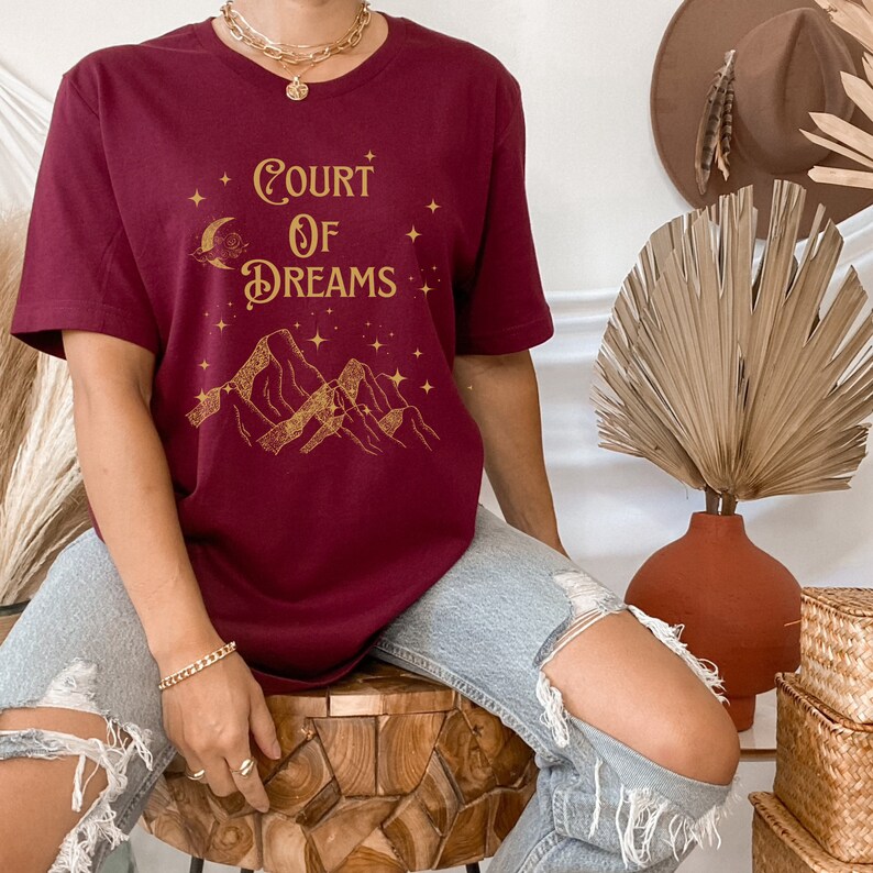 May include: A maroon t-shirt with a gold graphic design featuring mountains, stars, and the text "Court of Dreams".