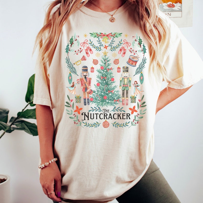 May include: Cream-colored t-shirt featuring a festive "Nutcracker" design. The graphic includes a Christmas tree, nutcracker figures, ballet shoes, and the text "The Nutcracker" in a decorative font. The design is surrounded by a wreath of leaves and ornaments.