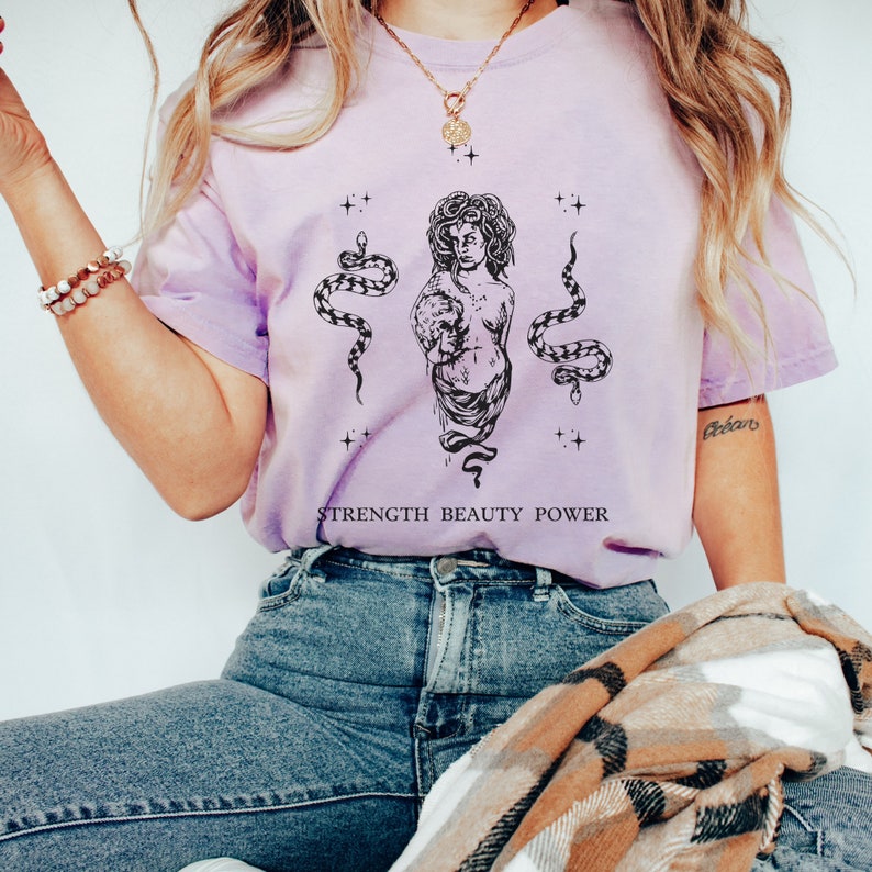 May include: A light purple t-shirt with a black and white graphic of a woman with snakes and the text "STRENGTH BEAUTY POWER".