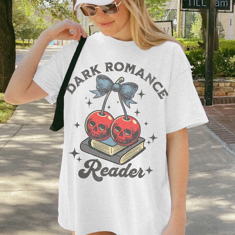 K&ouml;nnte beinhalten: Wei&szlig;es T-Shirt mit einem Grafikdesign mit zwei roten Kirschen mit Totenkopfgesichtern, einer blauen Schleife und dem Text "DARK ROMANCE Reader". Das Design ist auf ein wei&szlig;es, &uuml;bergro&szlig;es T-Shirt gedruckt.
