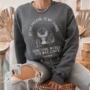 Op de afbeelding: Een donkergrijs sweatshirt met een witte grafische print van een hand die een kristallen bol vasthoudt met kaarsen en sterren. De tekst luidt "By the pricking of my thumbs, something wicked this way comes - Shakespeare".