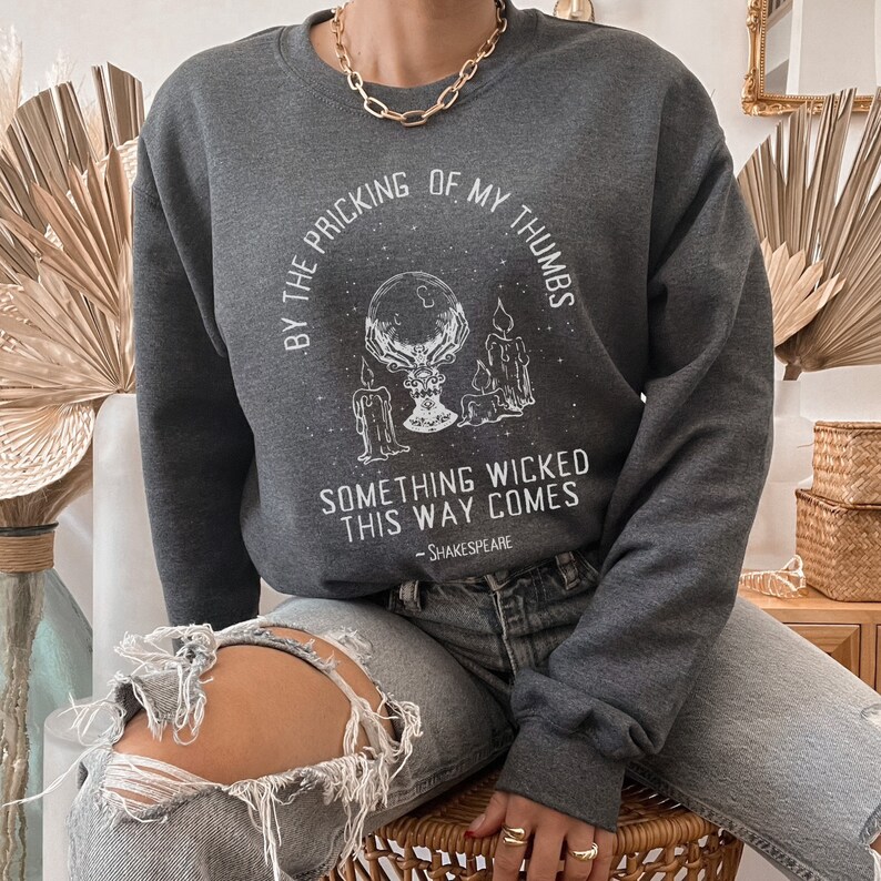 Op de afbeelding: Een donkergrijs sweatshirt met een witte grafische print van een hand die een kristallen bol vasthoudt met kaarsen en sterren. De tekst luidt "By the pricking of my thumbs, something wicked this way comes - Shakespeare".