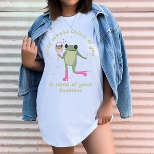 May include: White t-shirt with a green frog wearing pink shoes and holding a mirror. The text on the shirt reads "What others think of you is none of your business."