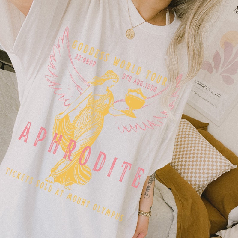 May include: White t-shirt with a pink and yellow graphic print of a woman with wings holding a cup. The text "GODDIES WORLD TOUR 22:00HR 5TH AUG. 1998" and "APHRODITE" is printed on the shirt. The text "TICKETS SOLD AT MOUNT OLYMPUS" is printed at the bottom of the shirt.