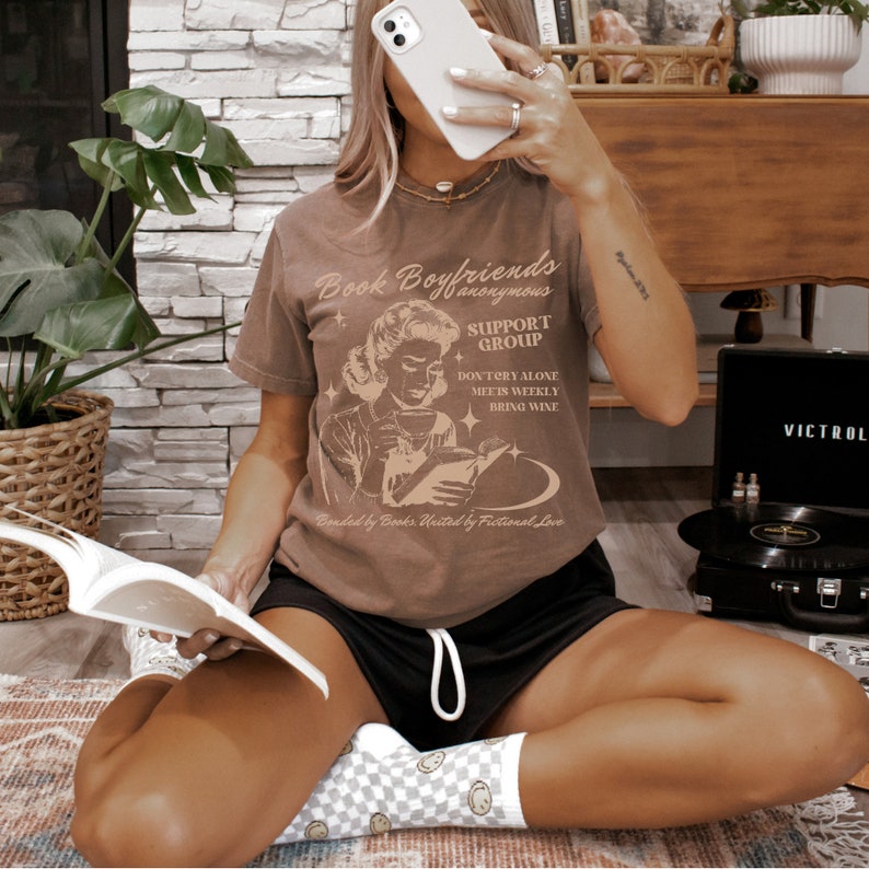May include: A brown t-shirt with a vintage-style illustration of a woman reading a book. The text on the shirt reads "Book Boyfriends anonymous SUPPORT GROUP DON'TERY ALONE MEETS WEEKLY BRING WINE Bonded by Books. United by Fictional Love".