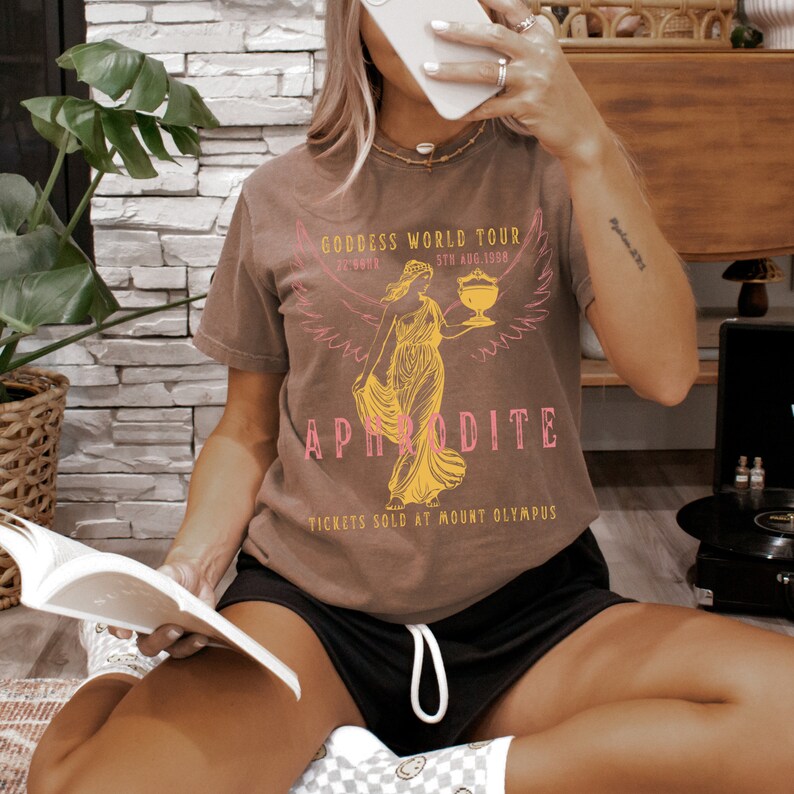 May include: A brown t-shirt with a graphic of a woman with wings holding a cup. The text on the shirt reads "Goddess World Tour", "Aphrodite", and "Tickets Sold at Mount Olympus".