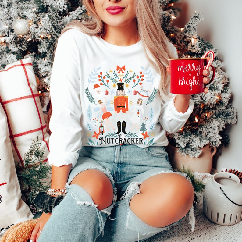 May include: White long-sleeve shirt with a colorful illustration of a Nutcracker and other characters from the ballet. The shirt has the text "The Nutcracker" printed on the bottom. The person is holding a red mug with the text "merry + bright".