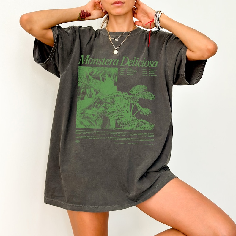 May include: A dark gray oversized t-shirt with a green graphic print of a Monstera Deliciosa plant. The shirt has a crew neck and short sleeves.