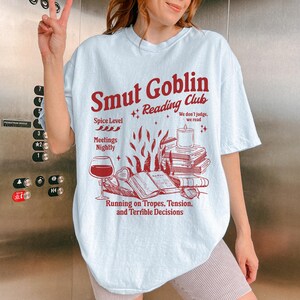 May include: Light blue t-shirt with a red graphic design that says "Smut Goblin Reading Club". The graphic includes a book, a wine glass, a candle, and the text "Spice Level", "Meetings Nightly", "We don't judge, we read", and "Running on Tropes, Tension, and Terrible Decisions".