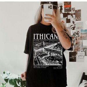 May include: Black t-shirt with a white graphic print of a bridge and the text "ITHICAN". The text below the graphic reads "Raging seas, hidden jungles and a bridge of secrets. Painted by King Area Keitell."