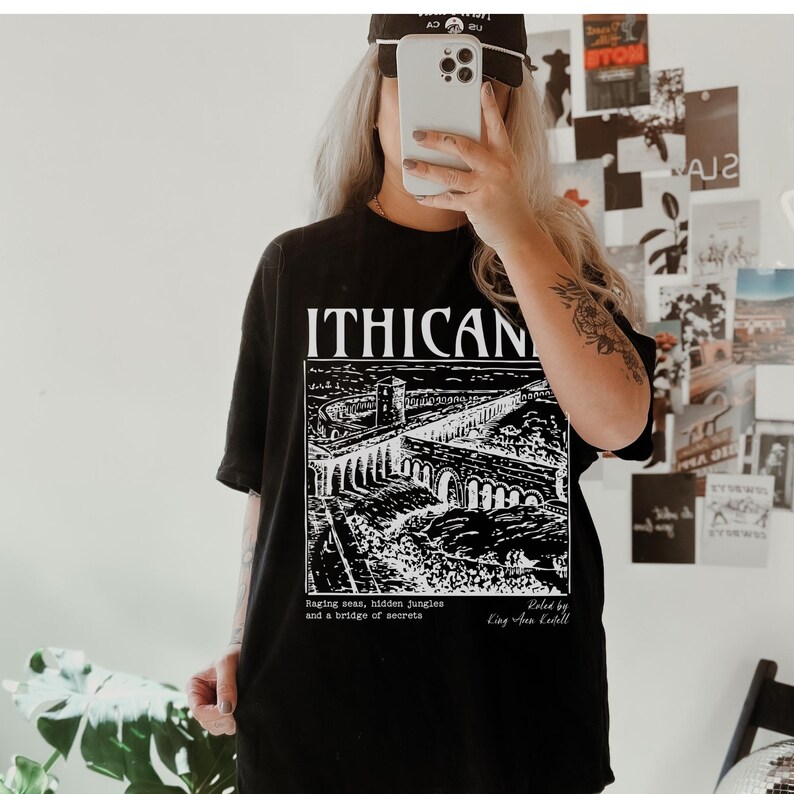May include: Black t-shirt with a white graphic print of a bridge and the text "ITHICAN". The text below the graphic reads "Raging seas, hidden jungles and a bridge of secrets. Painted by King Area Keitell."