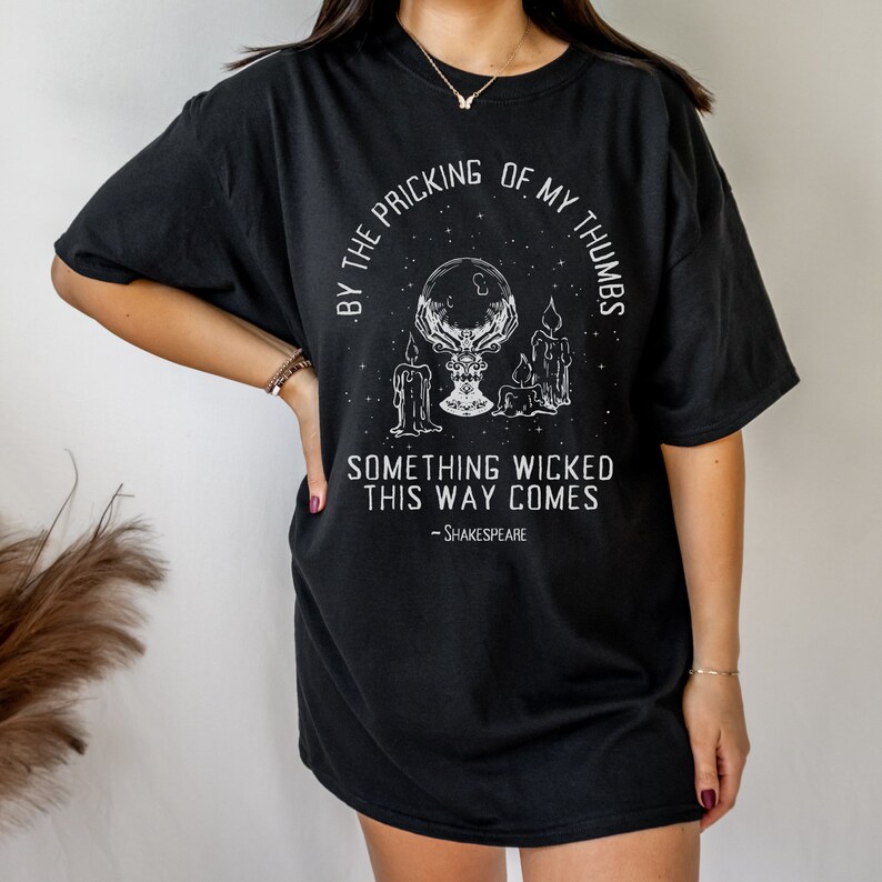 May include: Black oversized t-shirt with a white graphic print of a hand holding a crystal ball with candles and stars. The text reads "By the pricking of my thumbs, something wicked this way comes - Shakespeare."