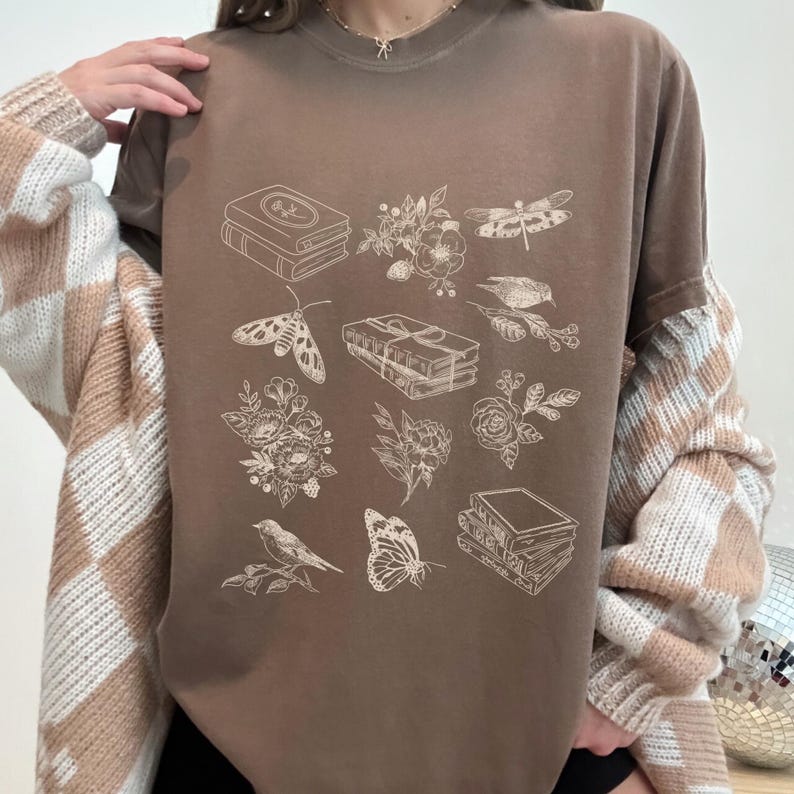 May include: A brown t-shirt with a white graphic print of various illustrations including books, flowers, butterflies, and dragonflies.