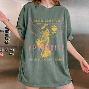 May include: A green t-shirt with a graphic print of a woman holding a cup. The text on the shirt reads "Goddess World Tour 5th Aug. 1998 22:69HR Aphrodite Tickets Sold at Mount Olympus".