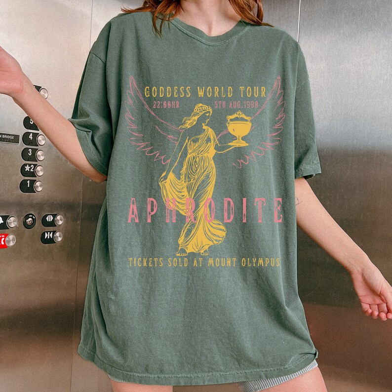 May include: A green t-shirt with a graphic print of a woman holding a cup. The text on the shirt reads "Goddess World Tour 5th Aug. 1998 22:69HR Aphrodite Tickets Sold at Mount Olympus".