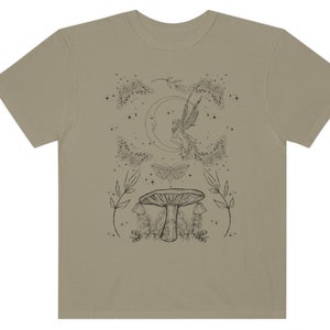 Pu&ograve; includere: Una t-shirt beige con un disegno a linea nera di una fata che vola vicino a una luna crescente, circondata da farfalle e funghi. Il design include anche stelle ed elementi floreali.