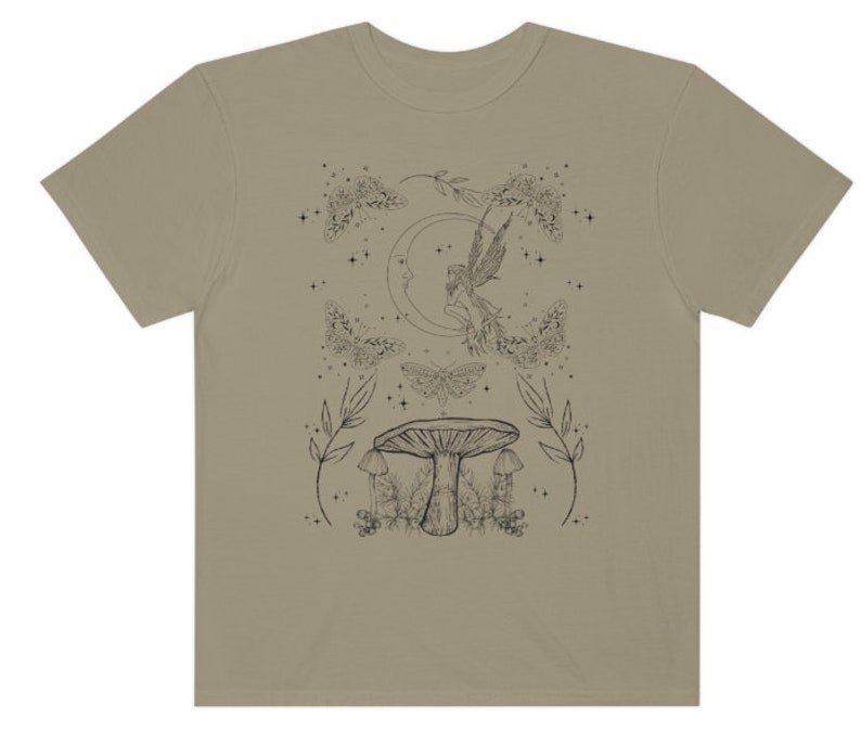 Pu&ograve; includere: Una t-shirt beige con un disegno a linea nera di una fata che vola vicino a una luna crescente, circondata da farfalle e funghi. Il design include anche stelle ed elementi floreali.