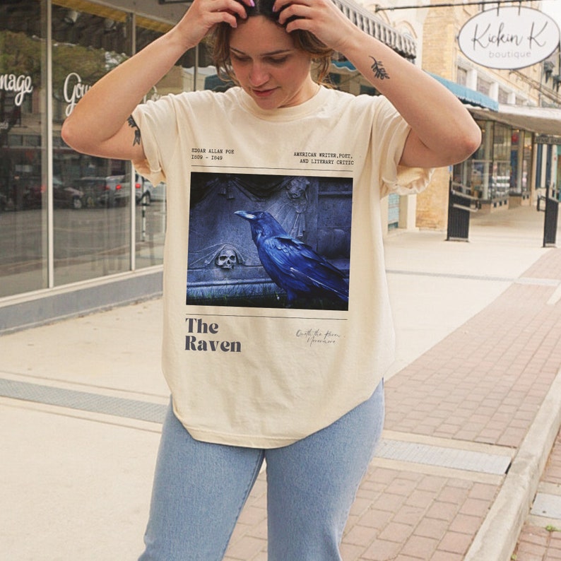 May include: A beige t-shirt with a graphic print of a raven perched on a tombstone. The text "The Raven" is printed below the image. The text "Edgar Allan Poe 1809 - 1849" is printed in the top left corner. The text "American Writer, Poet, and Literary Critic" is printed in the top right corner.