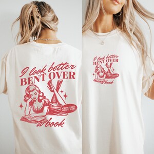 K&ouml;nnte beinhalten: Cremefarbenes T-Shirt mit roter Grafik. Das Design zeigt eine Vintage-Illustration einer lesenden Frau mit dem Text "I look better BENT OVER @book" und "CERTIFIED GOOD GIRL" auf Vorder- und R&uuml;ckseite.