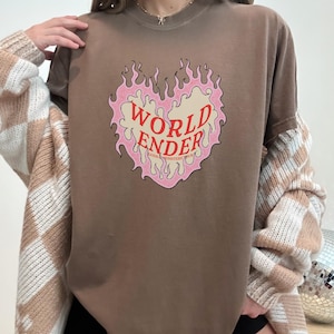 May include: A brown t-shirt with a pink and red graphic of a heart with flames. The text "WORLD ENDER" is inside the heart. The text "GODS & MONSTERS YOUTH" is below the heart.