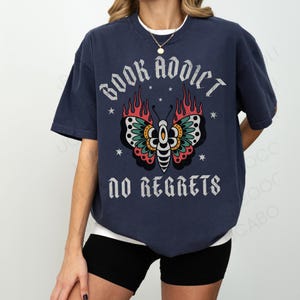 May include: Navy blue t-shirt with the text "BOOK ADDICT NO REGRETS" and a butterfly design. The butterfly has red, green, and yellow details with flames on the wings. The shirt is a relaxed fit.