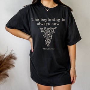 May include: Black oversized t-shirt featuring a quote from Mary Shelley: "The beginning is always now". The quote is printed in white with a decorative floral design.