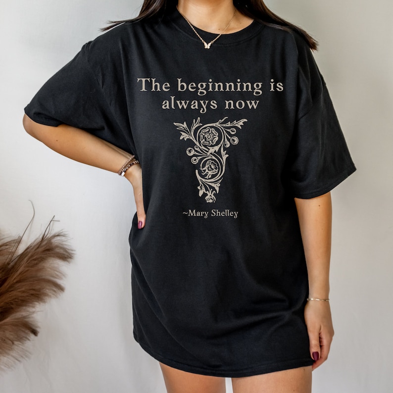 May include: Black oversized t-shirt featuring a quote from Mary Shelley: "The beginning is always now". The quote is printed in white with a decorative floral design.