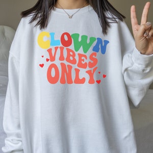 Może przedstawiać: Biała bluza z napisem "CLOWN VIBES ONLY" w kolorach tęczy z czerwonymi sercami.
