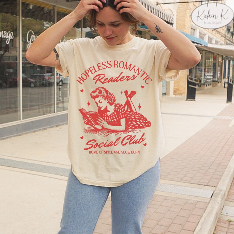 Peut inclure: Un t-shirt beige avec un motif rouge repr&eacute;sentant une femme lisant un livre. Le texte du motif est "Hopeless Romantic Readers Social Club Home of Spice and Slow Burn".