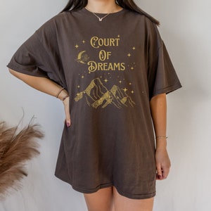 May include: An oversized brown t-shirt featuring a gold graphic print of mountains, a crescent moon, and stars. The text "Court of Dreams" is printed in gold on the shirt.