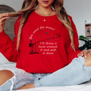 Pode incluir: Um moletom vermelho com a frase "You want the moon? I'll throw a lasso around it and pull it down" em texto branco. O desenho mostra uma pessoa laçando a lua. O moletom é de gola redonda e manga comprida.