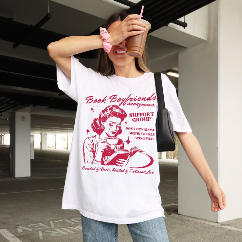 May include: White t-shirt with a red graphic of a woman reading a book and drinking coffee. The graphic says "Book Boyfriends Anonymous Support Group Don't Cry Alone Meets Weekly Bring Wine Bonded by Books United by Fictional Love."