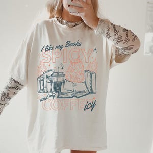 May include: Cream-colored oversized t-shirt with the text "I like my Books SPICY and my COFFEE icy" in teal and coral. The graphic includes books, a coffee cup, and flames. The shirt has long sleeves underneath.
