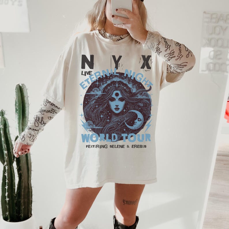 May include: Cream-coloured oversized t-shirt with a graphic featuring a woman's face within a blue circle, the text "NYX LIVE ETERNAL NIGHT WORLD TOUR" and "FEATURING SELENE & EREBUS". The shirt is worn over a patterned long-sleeved top.