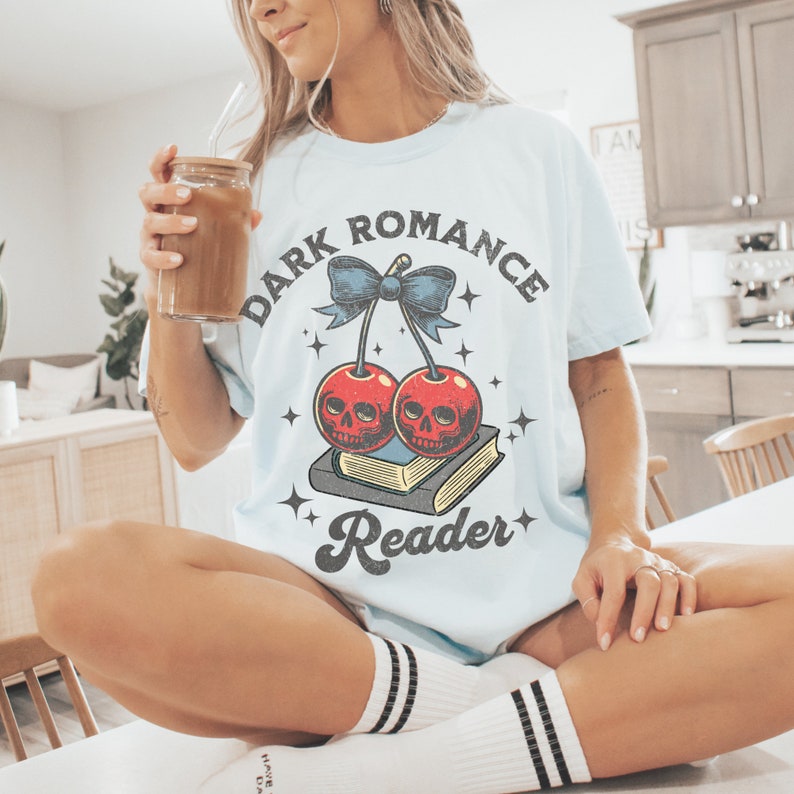 K&ouml;nnte beinhalten: Hellblaues T-Shirt mit dem Text "DARK ROMANCE READER" und einer Grafik von Kirschen mit Totenkopfgesichtern. Die Kirschen sind durch eine blaue Schleife verbunden und liegen auf einem Buch. Das Shirt hat eine lockere Passform.