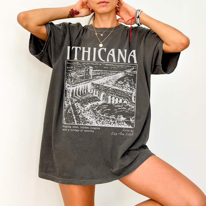 May include: A dark gray oversized t-shirt with a graphic print of a bridge and the text "ITHIČANA" in white. The text "Raging seas, hidden jungles and a bridge of secrets" and "Roled by King Aur Keitell" are also printed on the shirt.