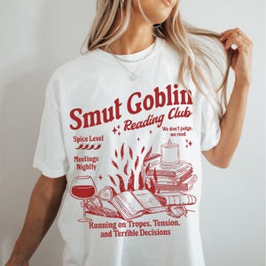 May include: White t-shirt with a red graphic design that says "Smut Goblin Reading Club". The graphic includes a stack of books, a lit candle, a glass of wine, and the text "Spice Level", "Meetings Nightly", "We don't judge, we read", and "Running on Tropes, Tension, and Terrible Decisions".