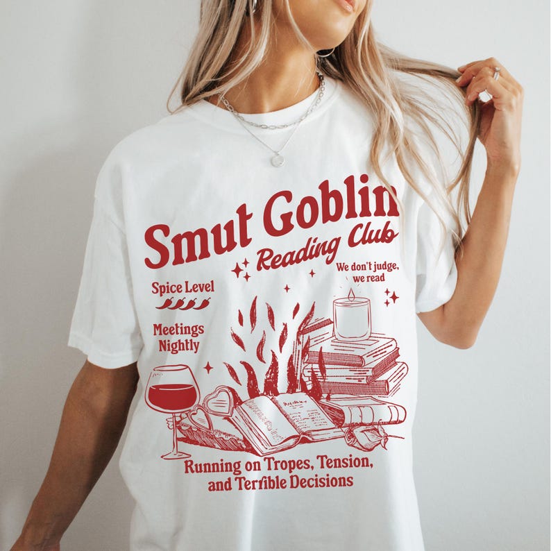 May include: White t-shirt with a red graphic design that says "Smut Goblin Reading Club". The graphic includes a stack of books, a lit candle, a glass of wine, and the text "Spice Level", "Meetings Nightly", "We don't judge, we read", and "Running on Tropes, Tension, and Terrible Decisions".