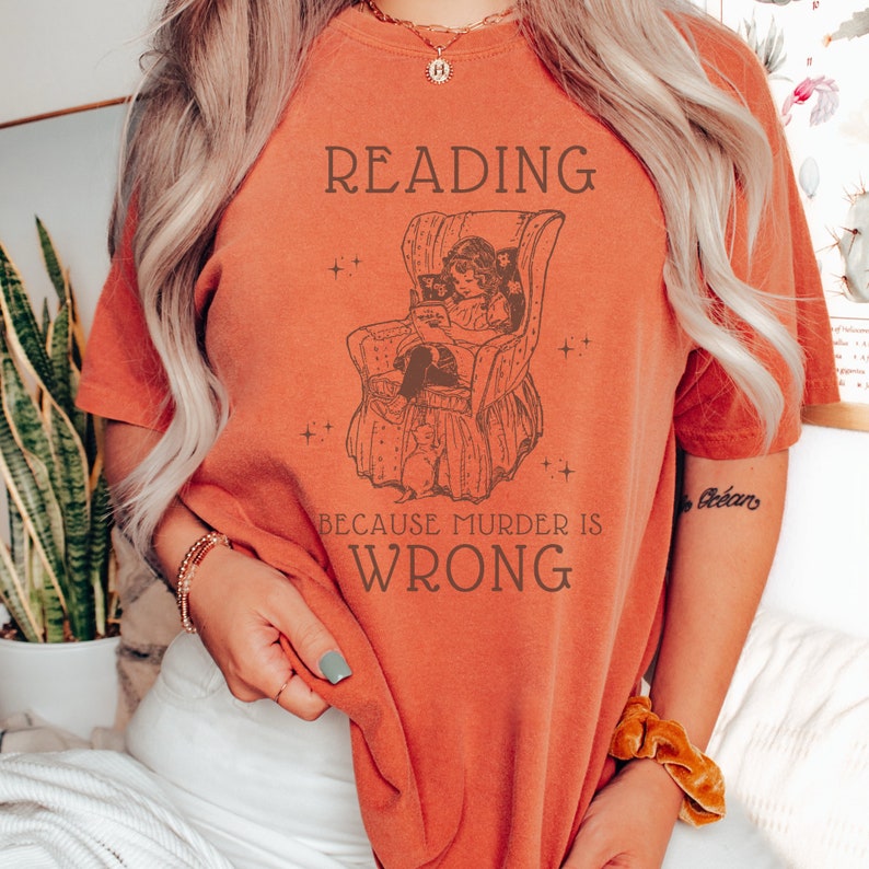 Puede incluir: Una camiseta naranja quemada con un gr&aacute;fico de una ni&ntilde;a leyendo en una silla. El texto en la camiseta dice "READING BECAUSE MURDER IS WRONG."