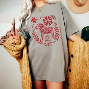 May include: A grey t-shirt featuring a red and white Scandinavian Dala horse design. The horse is surrounded by flowers and leaves.