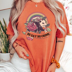 May include: Burnt orange t-shirt featuring a graphic of an opossum, roses, and a rainbow. The text "BE GAY DO CRIME" is displayed on a banner below the opossum. The shirt is a comfortable, casual style.