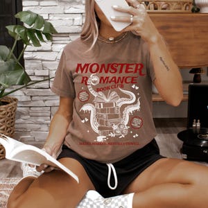 Pu&ograve; includere: T-shirt marrone con la scritta rossa "MONSTER ROMANCE BOOK CLUB". La grafica presenta una pila di libri con tentacoli e cuori. La frase "MATED, MARKED, MENTALLY UNWELL" &egrave; stampata sotto.
