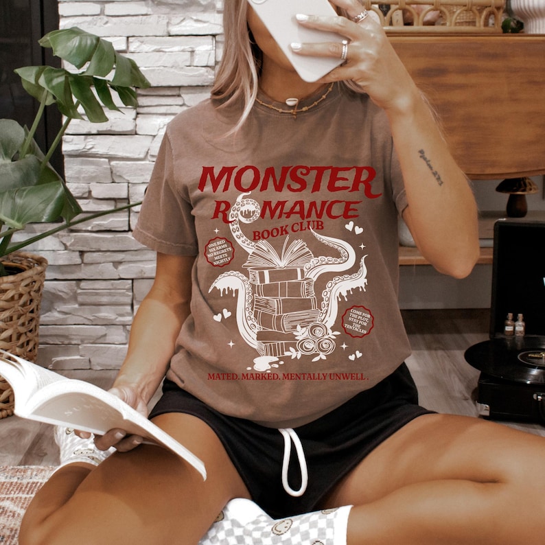 Pu&ograve; includere: T-shirt marrone con la scritta rossa "MONSTER ROMANCE BOOK CLUB". La grafica presenta una pila di libri con tentacoli e cuori. La frase "MATED, MARKED, MENTALLY UNWELL" &egrave; stampata sotto.
