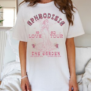 K&ouml;nnte beinhalten: Wei&szlig;es T-Shirt mit einem pinkfarbenen Grafikdesign, das eine Frau in einem flie&szlig;enden Kleid, eine Muschel und den Text "APHRODISIA 19 LOVE 62 TOUR THE GARDEN FRI AUGUST 5TH DOORS OPEN 8PM" zeigt.