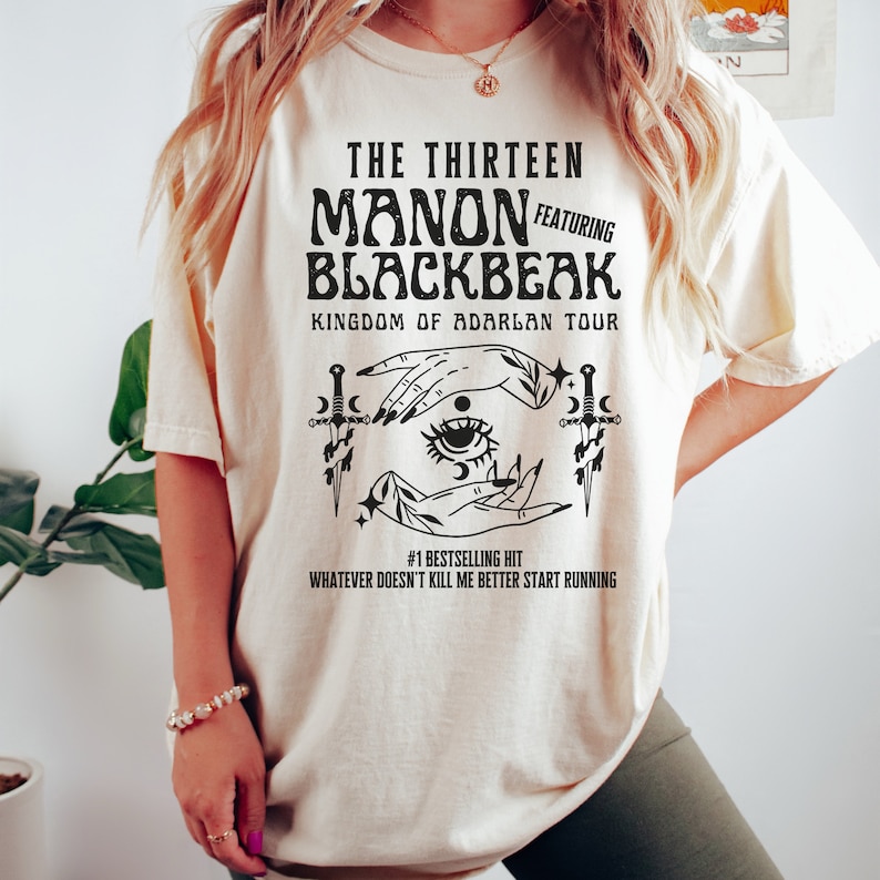 May include: A cream-coloured t-shirt with a black graphic design featuring the text "The Thirteen Manon Featuring Blackbeak Kingdom of Adarlan Tour" and a stylised illustration of a hand with an eye and daggers. The text "#1 Bestselling Hit Whatever Doesn't Kill Me Better Start Running" is also included in the design.