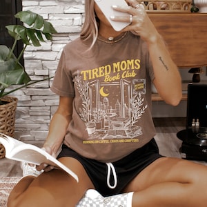 May include: A brown t-shirt featuring a graphic of a window with books, plants, and a city skyline. The text "Tired Moms Book Club" is printed in yellow above the window. The text "Running on coffee, chaos and chapters" is printed below the window.