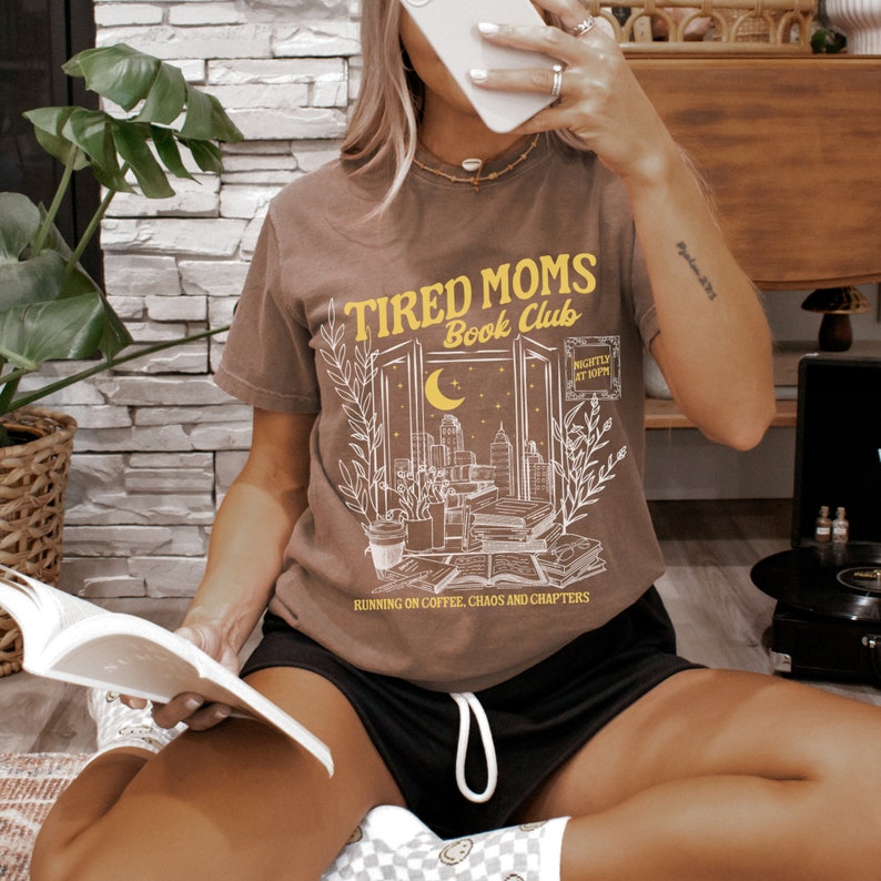 May include: A brown t-shirt featuring a graphic of a window with books, plants, and a city skyline. The text "Tired Moms Book Club" is printed in yellow above the window. The text "Running on coffee, chaos and chapters" is printed below the window.