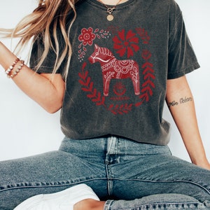 May include: A dark grey t-shirt featuring a red and white Scandinavian Dala horse design. The horse is surrounded by red floral designs.